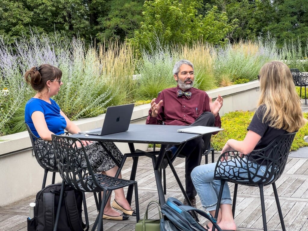 Three people sit around a table on an outdoor patio planted with many grasses and flowers.