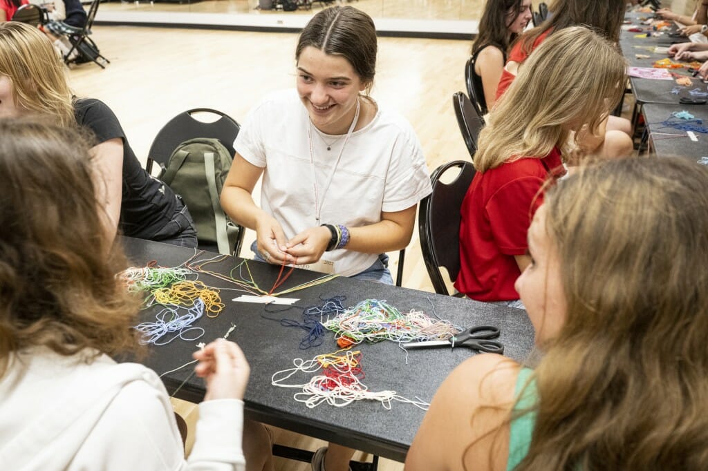 A student making a bracelet smiles at another student across the table.