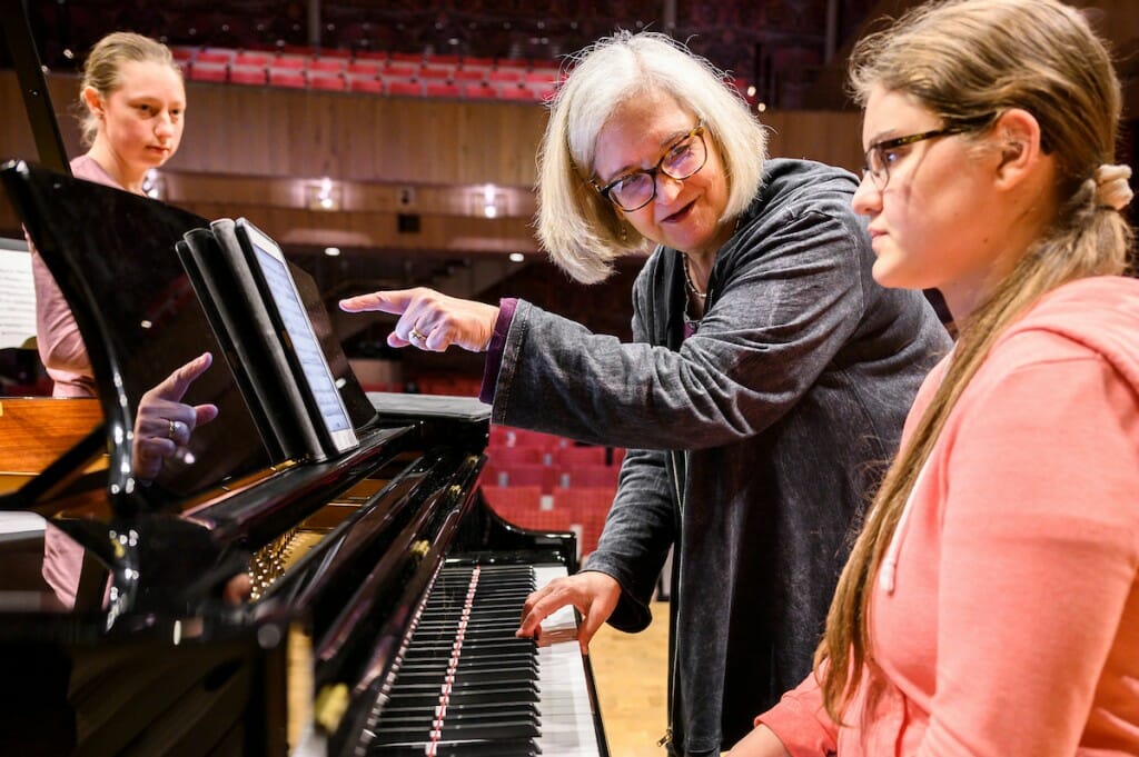 A person pointing at the sheet music on a piano while the piano player watches and listens