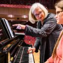 A person pointing at the sheet music on a piano while the piano player watches and listens