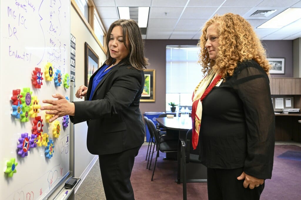 Two women look at a whiteboard with plastic gears on it, and talk.