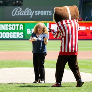 Chancellor Mnookin wears a navy blue Brewers shirt and points to Buck Badger as they both stand on the field at a Brewers game.