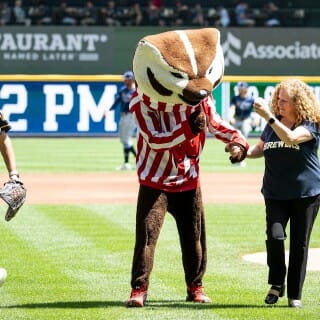 Chancellor Mnookin wears a navy blue Brewers shirt and walks hand-in-hand with Buck Badger as they leave the field during a Brewers game