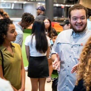 A young man in a blue shirt stands next to a young woman in a green shirt as he talks with Chancellor Mnookin, wearing a black shirt.