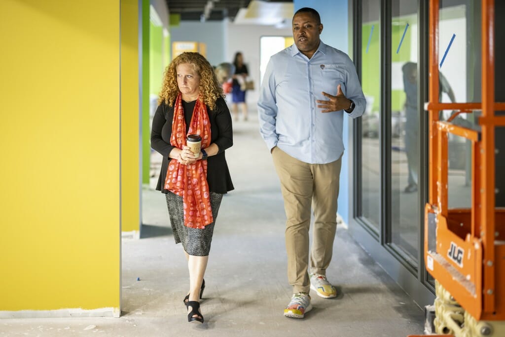 A woman in a bright red scarf and a man in a blue shirt and colorful sneakers walk side-by-side next to a bright yellow wall inside a building with construction equipment.