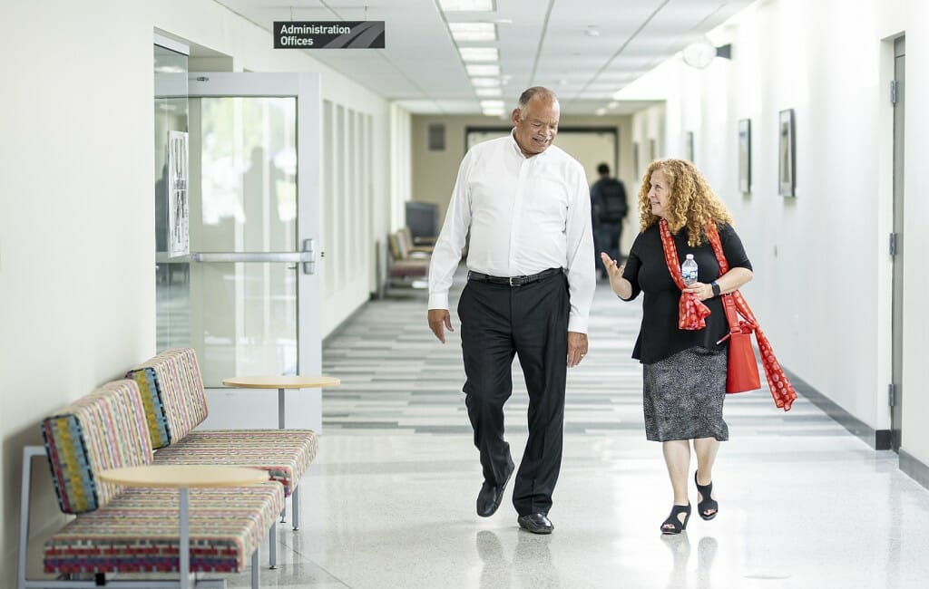 A man in a white shirt and black pants walks and talks along a hallway next to a woman in a black shirt and bright red scarf.