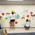 Four people apply paint to an intricate mural design on a wall.