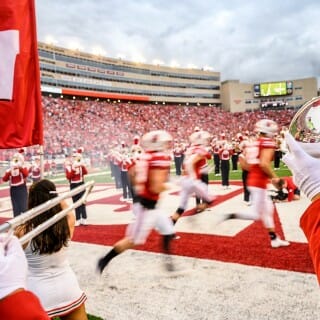 Badger Football players run through the endzone and onto the field, with cheerleaders cheering them.