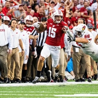 A football player runs down the sideline, with cheering coaches and fans visible in the background.