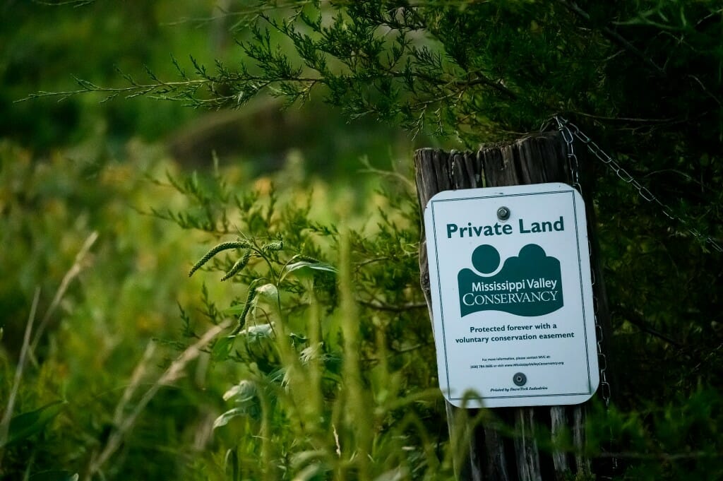 A white and green sign identifies private land that is part of the Mississippi Valley Conservancy. Yellow flowers are in the background.