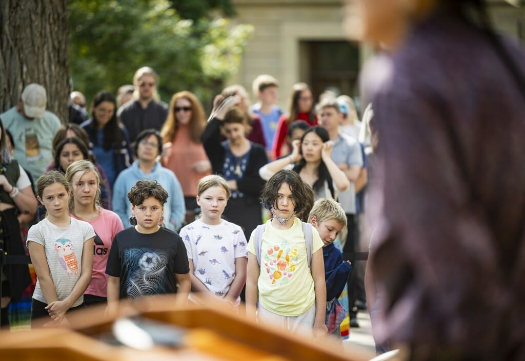 Children stand in several rows, looking up at a speaker.