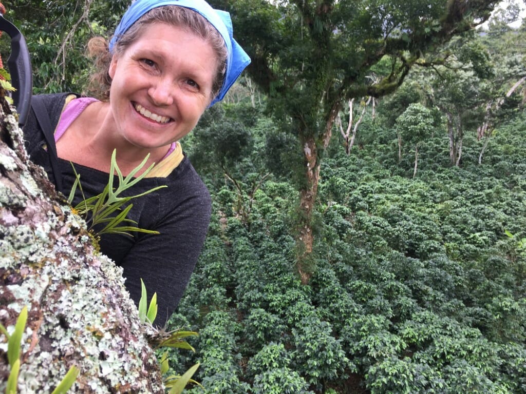 A woman smiles at the camera; behind her is a canopy of a tropical forest.