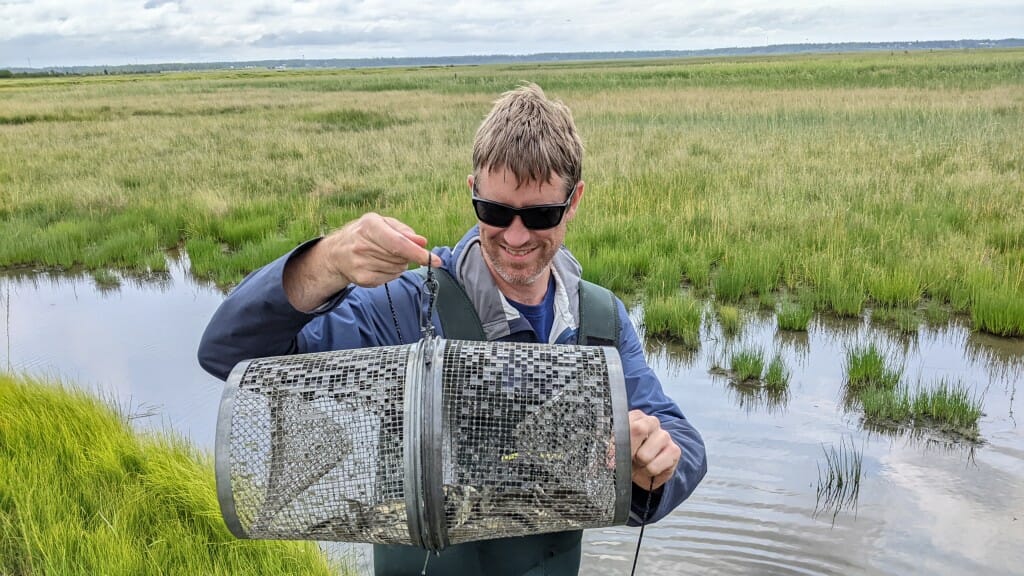 A man holds up a fish trap, in front of a river.