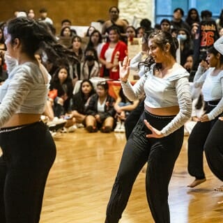 A group of seven dancers in powder blue tops and black pants move in unison with their right hands overhead and their left hands at their hips.