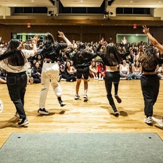 Five dancers in a V formation face away from the camera toward a seated, cheering audience.