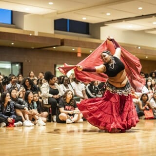 A belly dancer in a black top and read skirt leans forward and swings a red silk sheet overhead. A seated audience watches in the background.