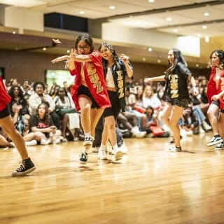 Six dancers wearing Sigma Psi Zeta sorority letter jackets dance in a line in front of a seated audience.