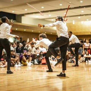 Five Black men in white shirts and black pants toss red and white walking sticks to each other while dancing for a seated audience.