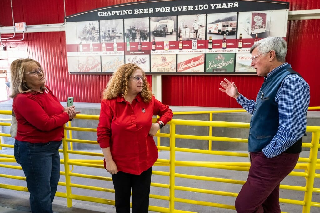 Kathy Bernier and Jennifer Mnookin listen to Dick Leinenkugel indoors. Behind them is a yellow metal gate against a red wall.