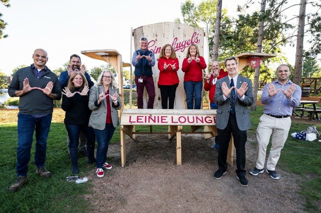 A group of people surround an oversized Adirondack chair. Three people stand on the chair. Everyone makes a letter W sign with their hands and smiles to the camera.