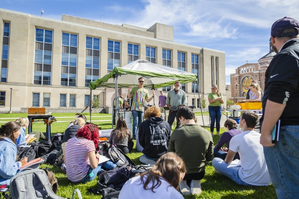 Joshua Calhoun stands in front of Memorial Library and a green and speaks to a group of students seated on the grass.