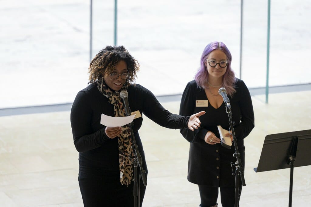 Two women stand in a museum, with a window in the background.