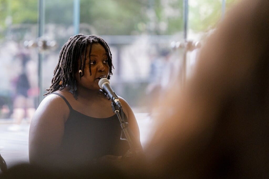 A woman speaks at a microphone, as onlookers listen.