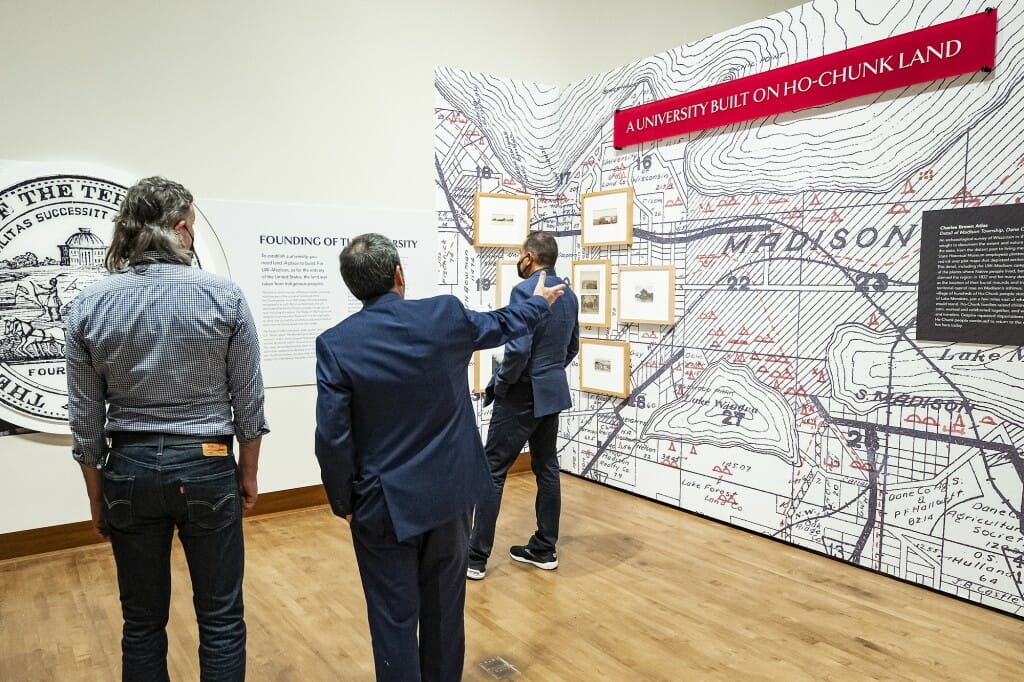 Three men face away from the camera toward an exhibit wall with a banner reading "A university built on Oh-Chunk land."