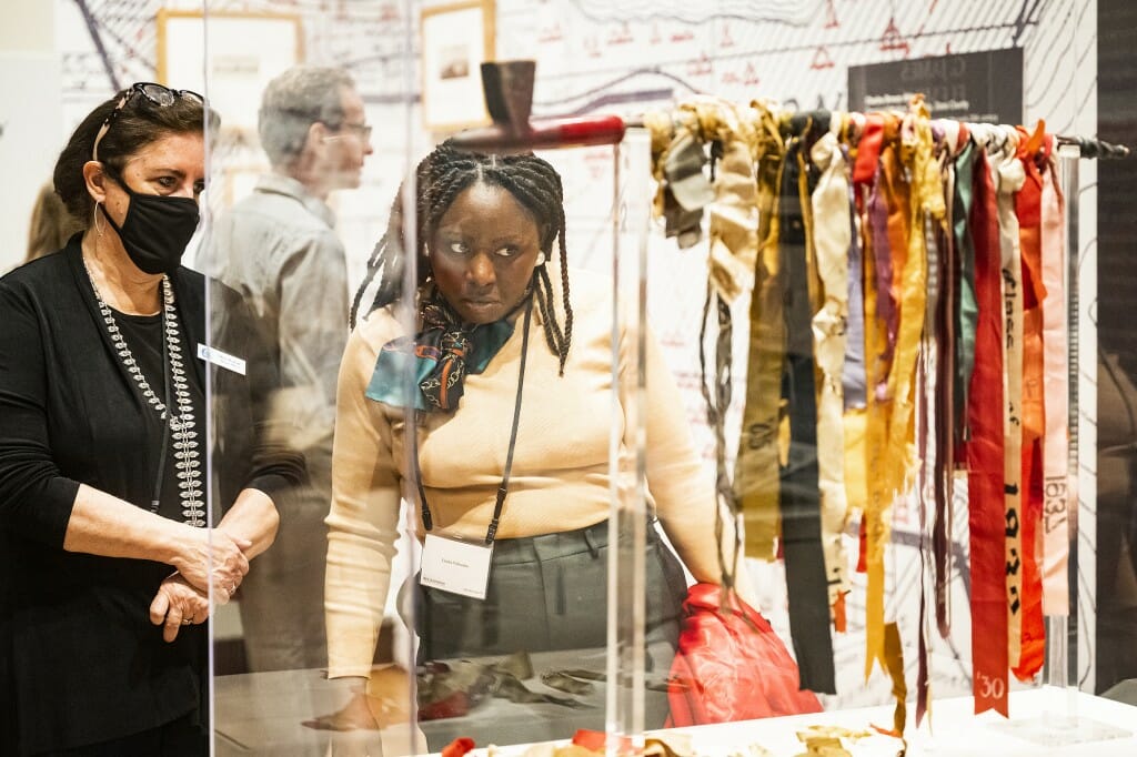 Two women stand behind a glass display case that holds a pipe decorated in long, colorful ribbons.