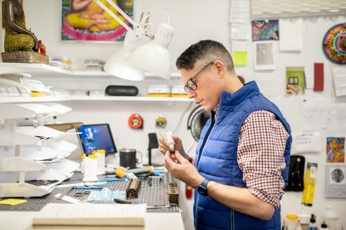 Michael Velliquette stands in an art studio at an elevated table with paper cutting supplies. He holds a small, white bottle of glue in one hand and a piece of paper in the other.