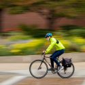 A cyclist wearing a neon jacket and black pants bikes in light rain past a flowerbed and a red brick building