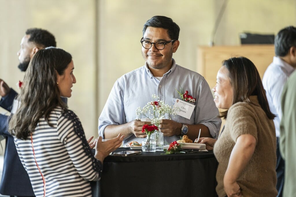 Three people, including Chris Saldaña (center), stand in conversation around a cocktail table in a brightly sunlit room.