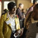 Angela Byars-Winston smiles in conversation. She wears a yellow leather jacket and white shirt and a corsage of red and white flowers.