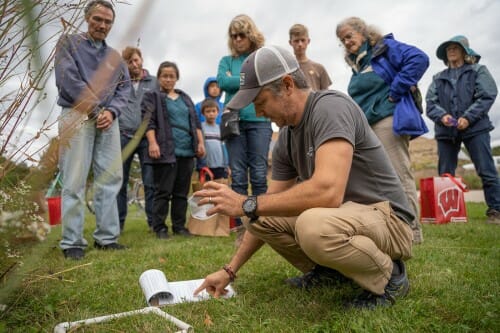 A man crouches over a clipboard laid on a lawn. A crowd of people stand behind him looking on.