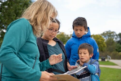 A woman holds a clipboard and talks with three people who look down at the clipboard. They are outdoors on a grass lawn.