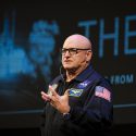 A close shot of Scott Kelly wearing his NASA flight suit in front of a dark stage background.