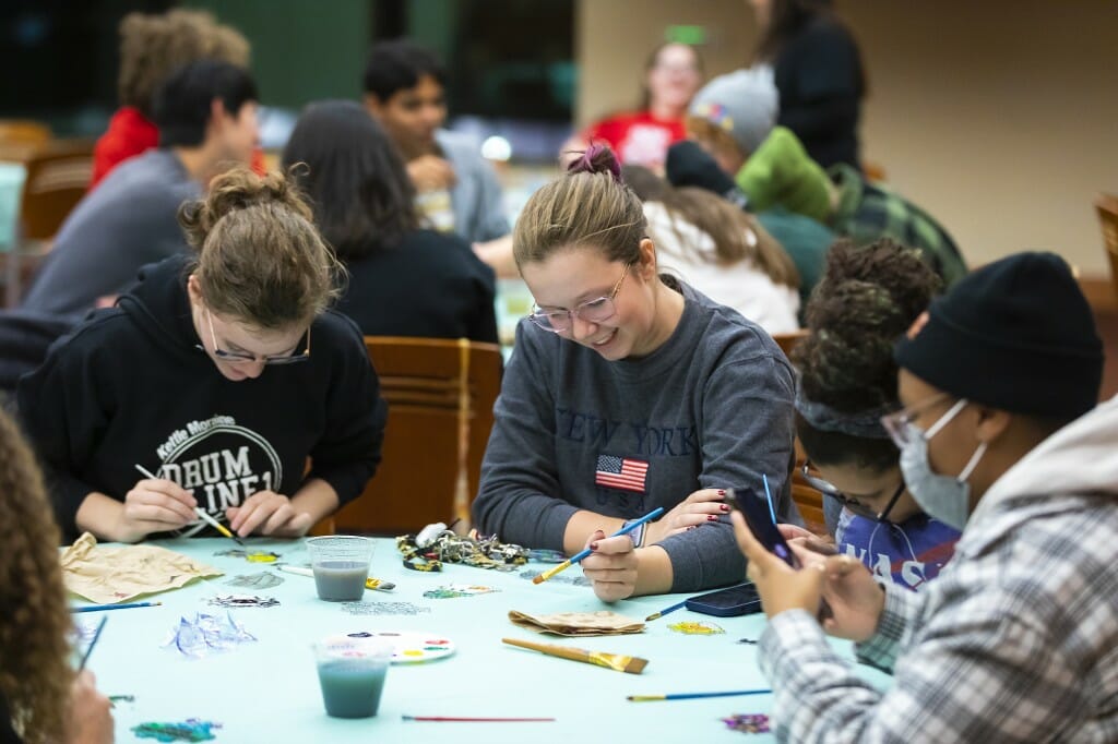 Two women sitting at a table smile as they paint.