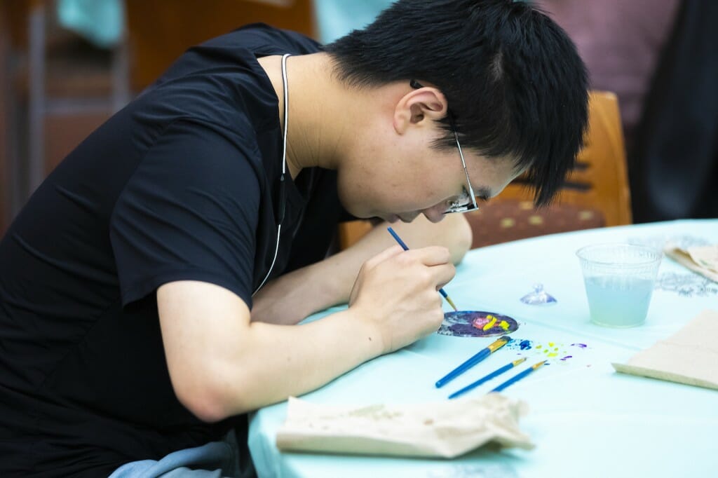 A man leans over to look closely at the suncatcher he's painting while sitting at a table.