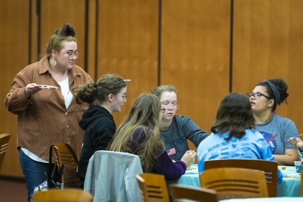 A woman stands near a table, handing paint to people sitting at the table.