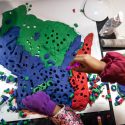 An overhead view of a table top covered in craft supplies. In the center, childrens' hands reach over a playdough sculpture of the state of Wisconsin.