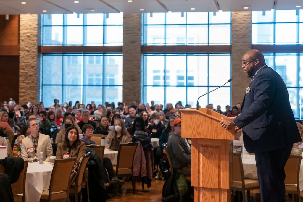 A man stands at a podium in front of a large crowd, backlit by large windows.