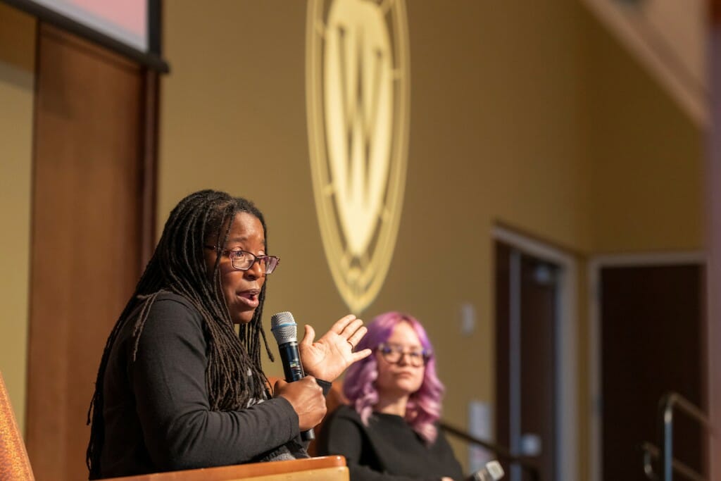 A woman holds a microphone and gesticulates, with a W crest representing the University of Wisconsin in the background.