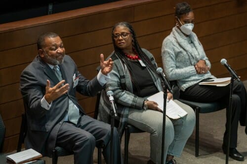 Three people sit in chairs, two listen while the other speaks into a microphone and gesticulates.