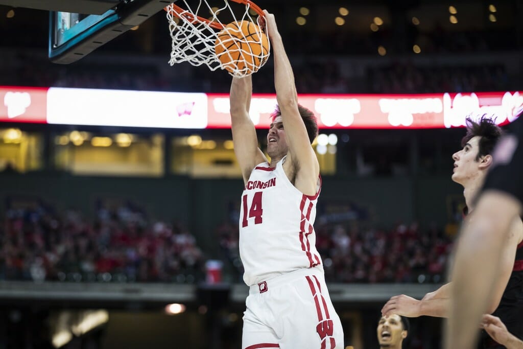 A man in a white Wisconsin basketball uniform holds onto the rim after dunking.