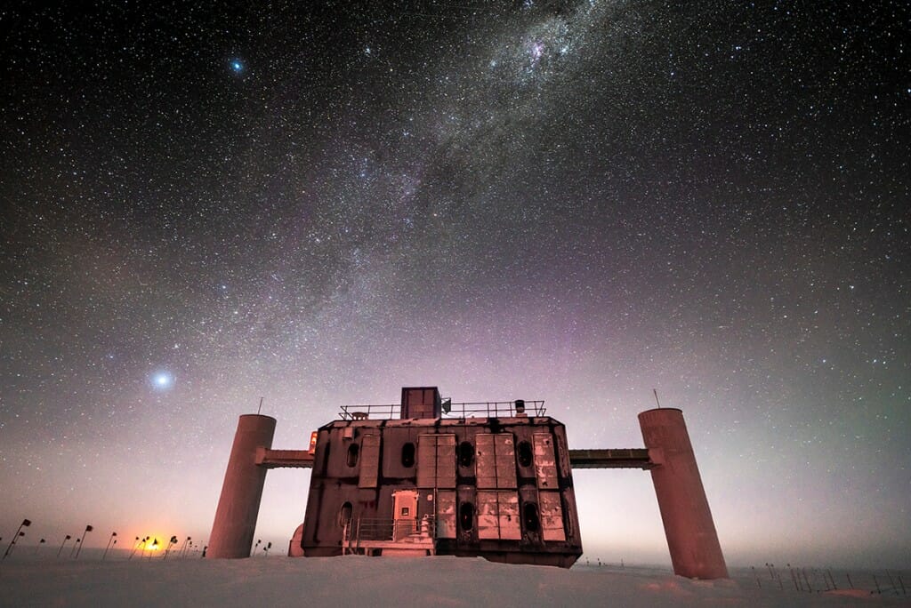 Front view of the IceCube Lab at twilight, with a starry sky showing a glimpse of the Milky Way overhead and sunlight lingering on the horizon.