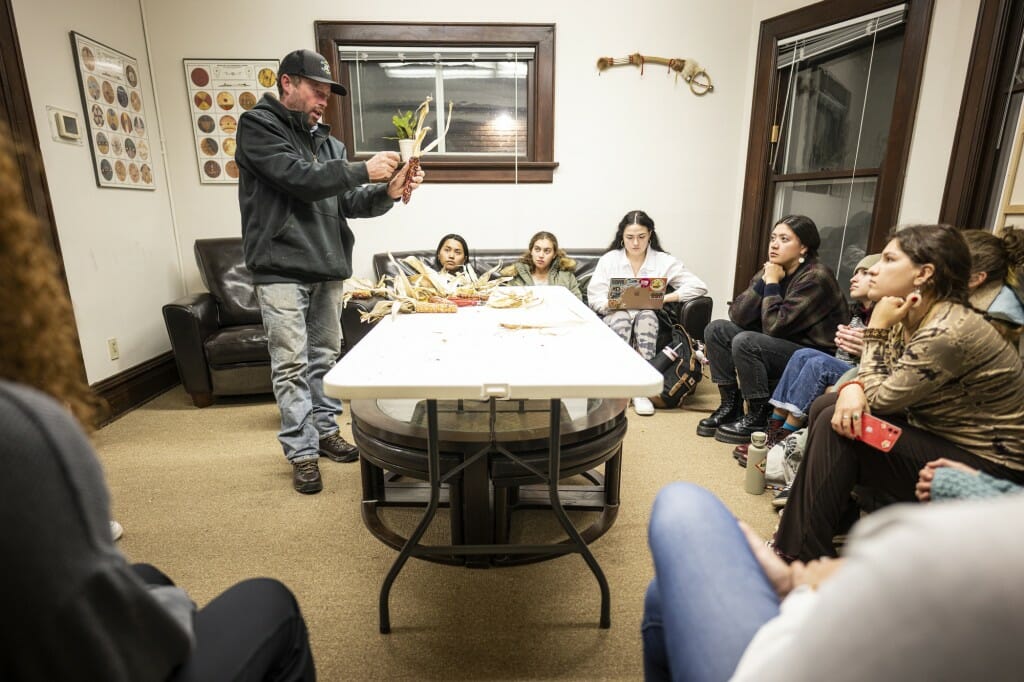 A man holds up corn cobs and braids him together as other people sit in a circle around him, watching.