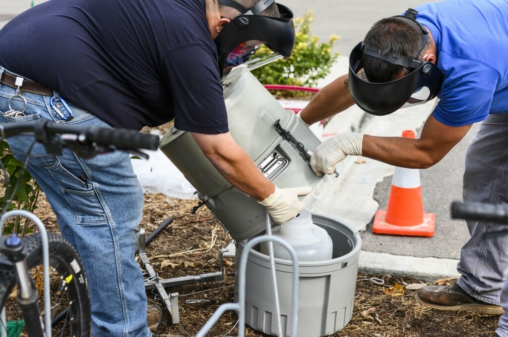 Wearing face shields, Physical Plant plumbers Jay Maier, left, and Adam Kundert hoist a compositing wastewater sampler from a sewer opening