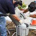 Wearing face shields, Physical Plant plumbers Jay Maier, left, and Adam Kundert hoist a compositing wastewater sampler from a sewer opening