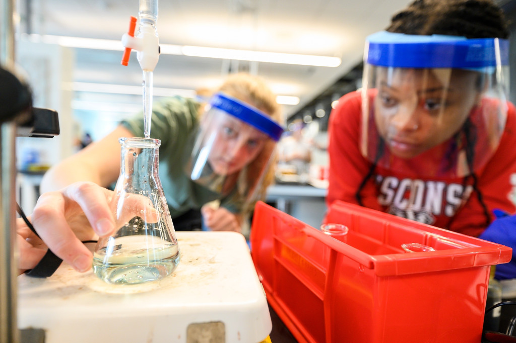 In a chemistry lab class, two students wearing face shields lean in to watch the liquid in a beaker change from blue to yellow.