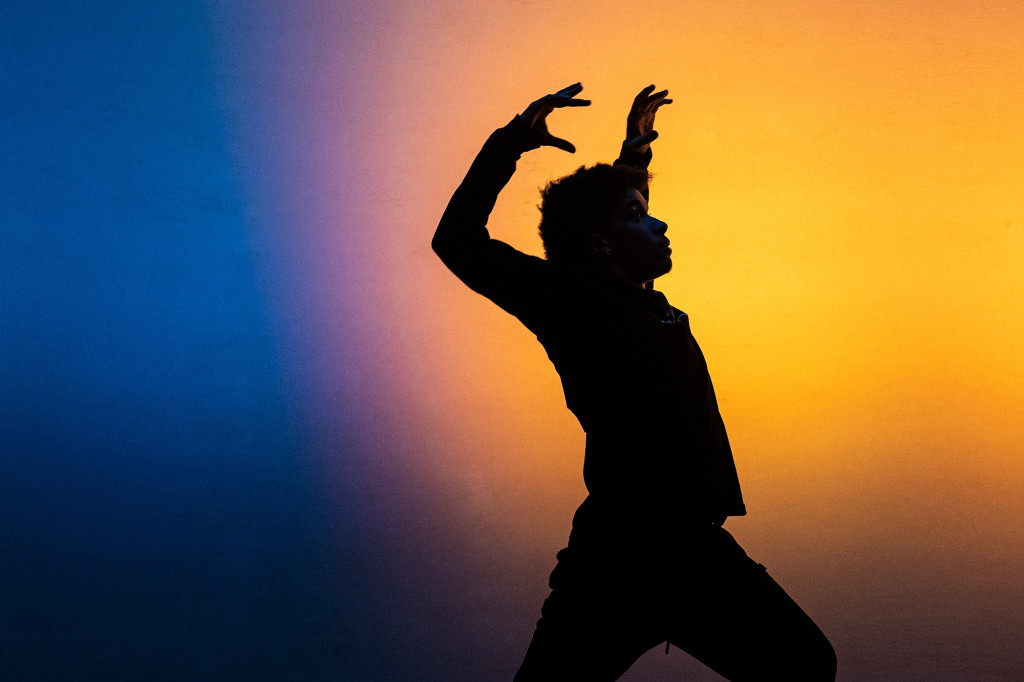 A dancer in silhouette raises his hands and arches his back in a full-body wave motion. He is backlit with blue and yellow light.